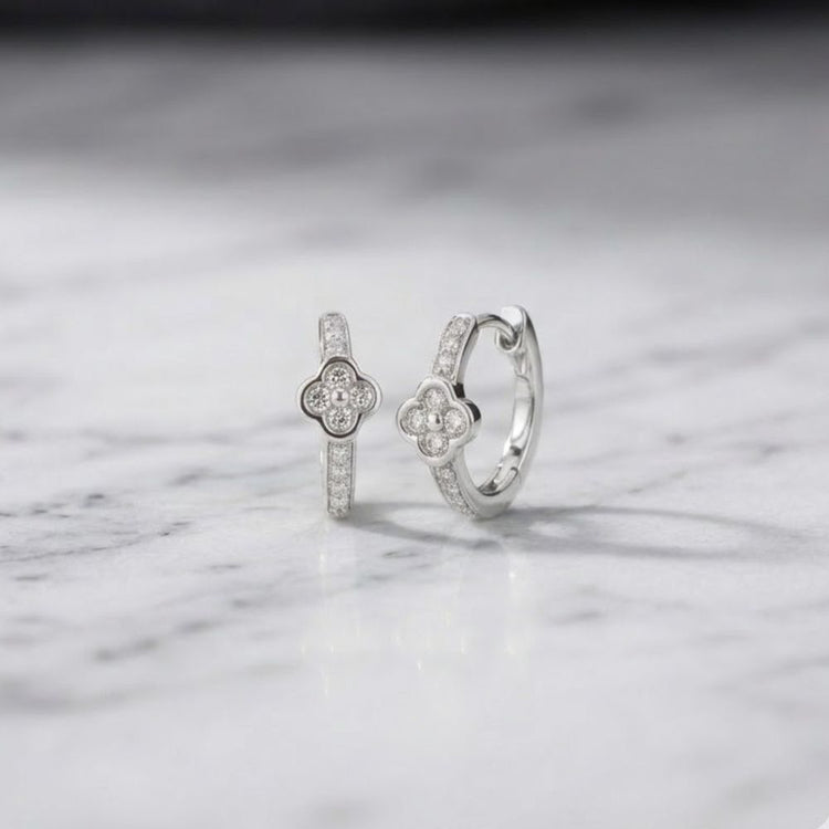 Two 1/10 CTW natural diamond clover hoop earrings, crafted in silver and featuring flower-shaped center designs, are elegantly displayed on a white marble surface.