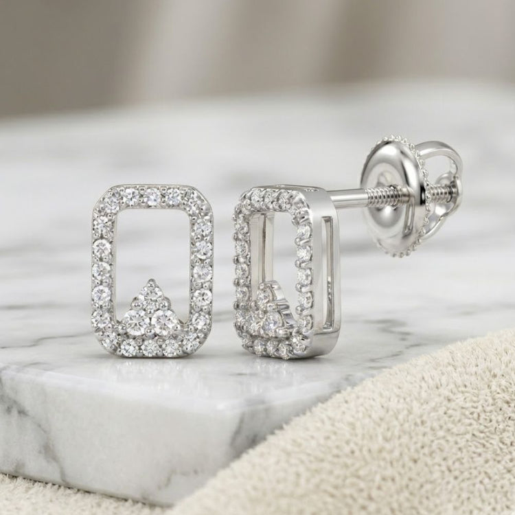 A pair of 1/6CTW natural diamond earrings in 10K gold with a rectangular shape, rounded corners, and an arrow-like accent, shown on marble with a beige cloth nearby.