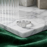 The 14k Gold Princess Diamond Halo Engagement Ring, featuring a stunning square halo setting, rests gracefully on a white marble tray as its band shimmers in the light. Soft green fabric and refined glass accents provide an elegant backdrop for this exquisite engagement ring.