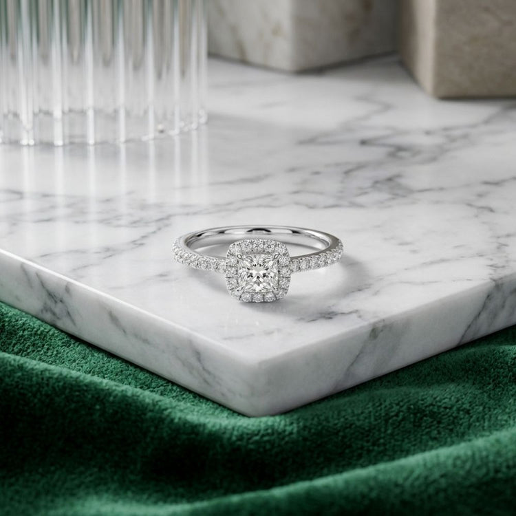 The 14k Gold Princess Diamond Halo Engagement Ring, featuring a stunning square halo setting, rests gracefully on a white marble tray as its band shimmers in the light. Soft green fabric and refined glass accents provide an elegant backdrop for this exquisite engagement ring.