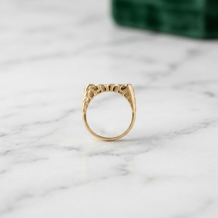 The 10k Yellow Gold Round Diamond Nugget Fashion Ring 1/2 Cttw is displayed upright on a white marble surface, showcasing its intricate sculpted design, with a blurred green object in the background.