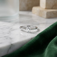 A 1/4CT Diamond Emerald Solitaire Ring in 14K Gold is displayed on a white marble surface beside a green velvet cloth and beige stone blocks in the background.
