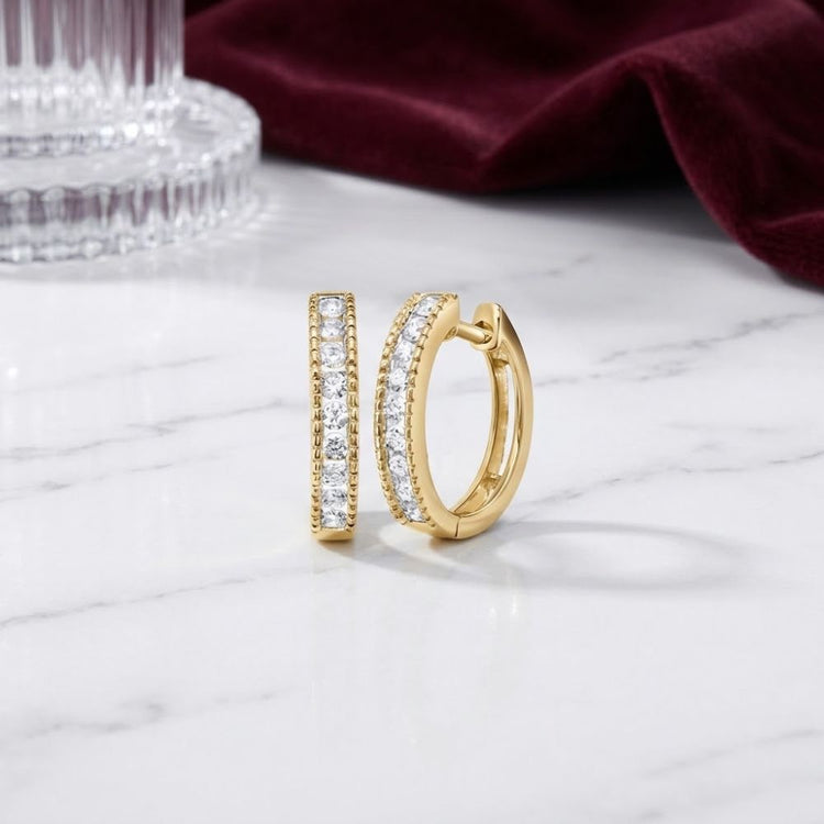 The 10K Gold Round Diamond Hoop Earrings 1/4 CTW, featuring two rows of small, sparkling diamonds set in elegant hoops, are showcased on a white marble surface. Their brilliance catches the light beautifully, with a glass vessel and deep red velvet fabric softly blurred in the background.