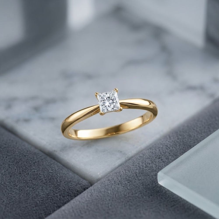 A beautiful 1/4CT Princess Diamond Solitaire Ring in 14K Gold is displayed on a marble and gray geometric background.