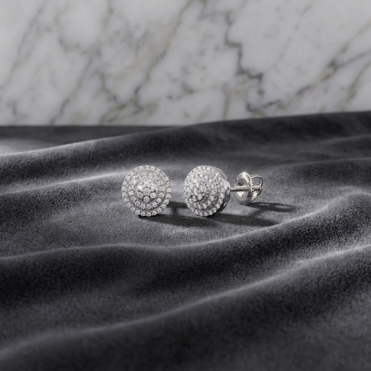 The 1/4 CTW Natural Diamond Round Earrings in 10K gold, each featuring concentric circles of sparkling natural diamonds, are displayed on a soft gray fabric with a light marble background.