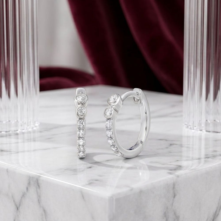 Two 1/10 CTW Natural Diamond Hoop Earrings crafted in 10K gold are displayed on a white marble surface, with clear glass columns and a burgundy velvet curtain in the background.
