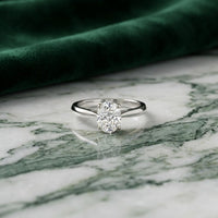 A 1/4 CT Diamond Oval Solitaire Ring in 14K Gold sits gracefully on a green and white marble surface, complemented by a piece of dark green velvet fabric in the background.