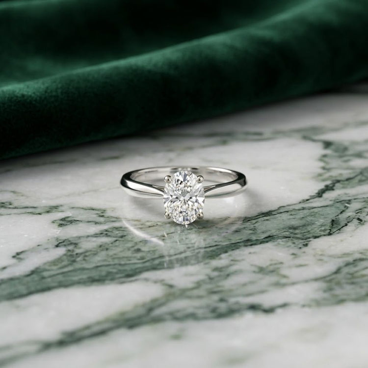 A 1/4 CT Diamond Oval Solitaire Ring in 14K Gold sits gracefully on a green and white marble surface, complemented by a piece of dark green velvet fabric in the background.