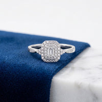 The 1/3CTW Diamond Emerald Double Halo Engagement Ring in 14k Gold, featuring a rectangular natural diamond surrounded by smaller diamonds, is displayed on a blue velvet and white marble surface.