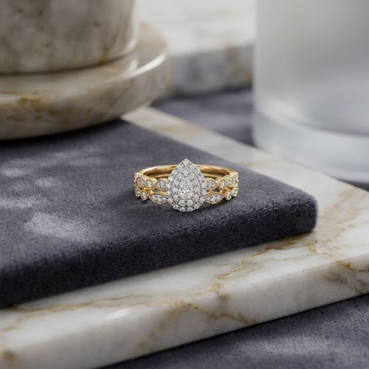 The 10k Gold Pear Diamond Halo Bridal Ring Set 3/8 Cttw features a striking teardrop-shaped diamond cluster surrounded by a halo of smaller diamonds, elegantly displayed on a gray velvet surface beside marble trays and a glass—an ideal choice for those seeking an elegant bridal wedding ring set.