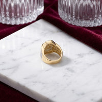A 10k Gold 7/8 CTTW Diamond Cross Men’s Ring rests on a white marble surface, surrounded by two crystal glasses and a burgundy velvet fabric.