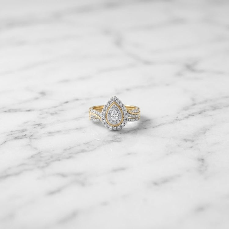 The 1/2 CTW Natural Diamond Pear Double Halo Bridal Set, crafted in 14K gold with a large teardrop-shaped diamond surrounded by a shimmering double halo of smaller diamonds, is elegantly displayed on a white marble surface.