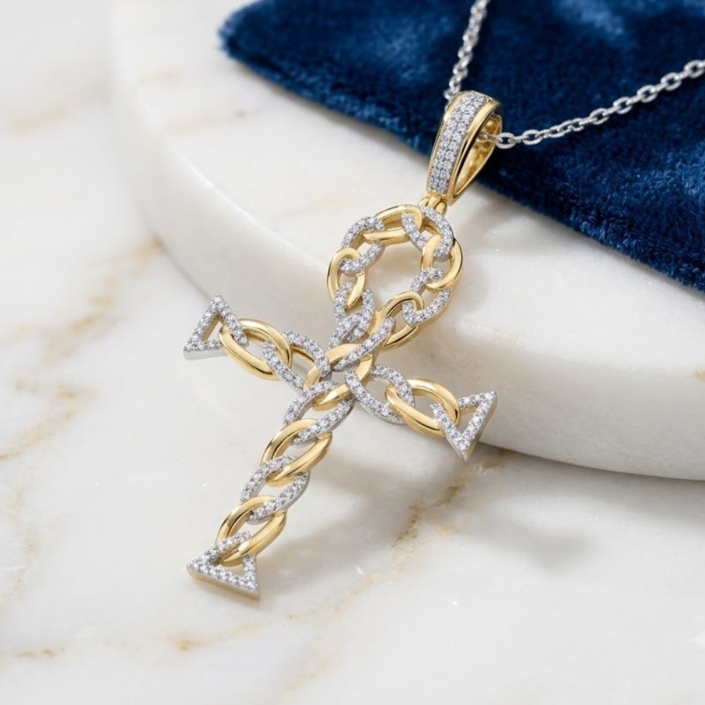 The 10K Gold Diamond Ankh Cross Pendant 3/8 CTW, crafted from yellow gold with braided details and sparkling diamonds, rests on a marble surface beside a blue velvet cloth.