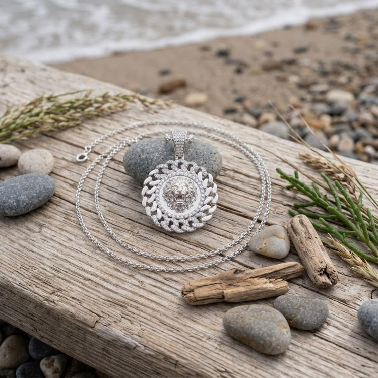A Diamond Lion Face Cuban Charm 1/2CT Sterling Silver men’s pendant necklace with a round, ornate lion face design rests on driftwood, surrounded by smooth stones, twigs, and sand near the beach, with ocean waves in the background.