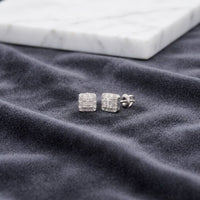 A pair of 1/6 CTW Natural Diamond Baguette Earrings rests on a soft, dark gray fabric with a white marble slab in the background.