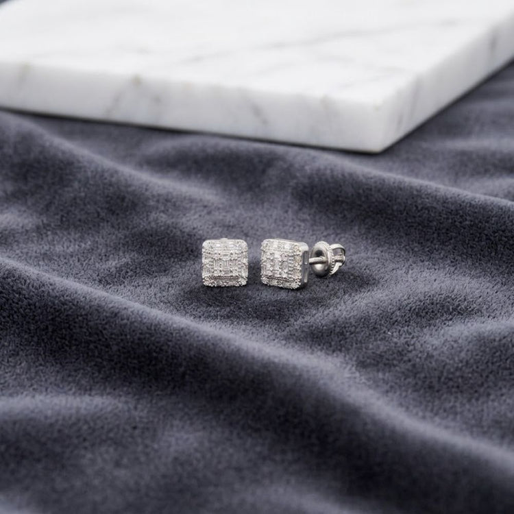 A pair of 1/6 CTW Natural Diamond Baguette Earrings rests on a soft, dark gray fabric with a white marble slab in the background.