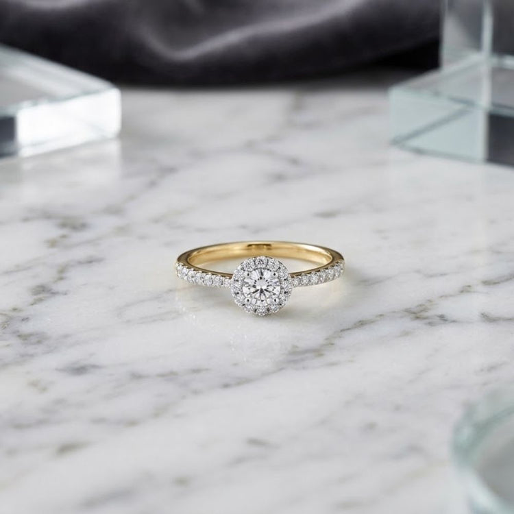 The 14k Gold Round Diamond Halo Engagement Ring is showcased on a white marble surface featuring delicate grey veining, with glass blocks and a soft, dark fabric accenting the background.