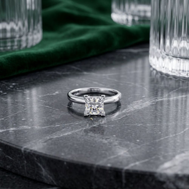 A 14k White Gold Princess Diamond Bridal Ring 3/4 Cttw gleams atop a polished dark marble surface. Clear glass tumblers and a green velvet cloth in the background accentuate the timeless elegance of its princess-cut diamond and refined setting.