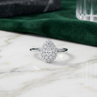 Introducing the 14k Gold Pear Diamond Halo Engagement Ring, featuring a captivating teardrop-shaped center diamond encircled by a sparkling halo of smaller diamonds, all set in luxurious 14k gold. This exquisite engagement ring is beautifully showcased on a marble surface, complemented by deep green fabric and delicate glass accents in the background.