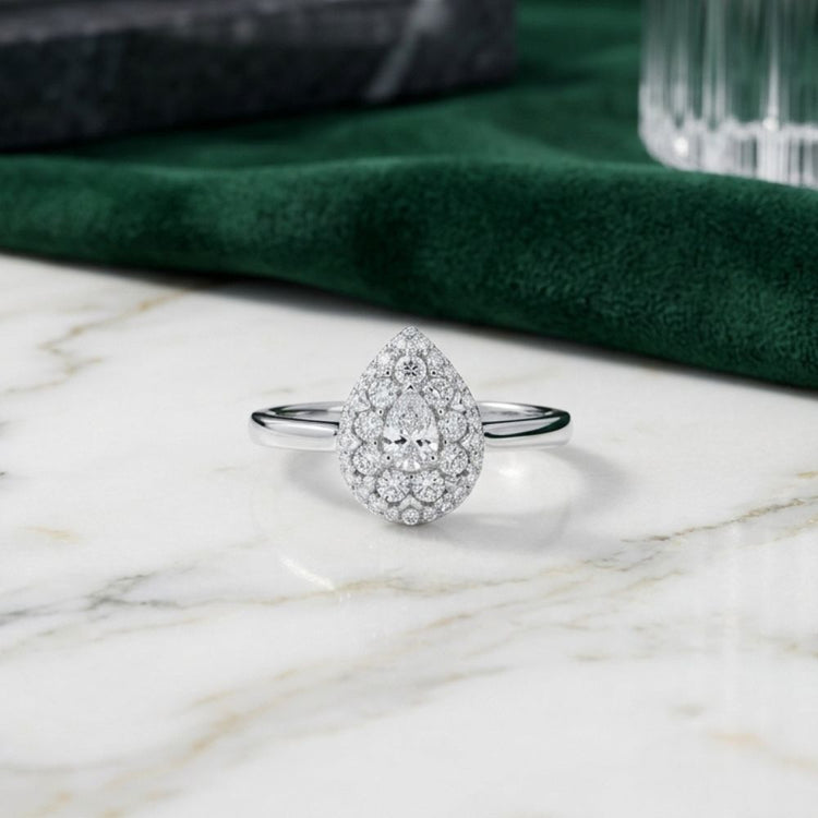 Introducing the 14k Gold Pear Diamond Halo Engagement Ring, featuring a captivating teardrop-shaped center diamond encircled by a sparkling halo of smaller diamonds, all set in luxurious 14k gold. This exquisite engagement ring is beautifully showcased on a marble surface, complemented by deep green fabric and delicate glass accents in the background.