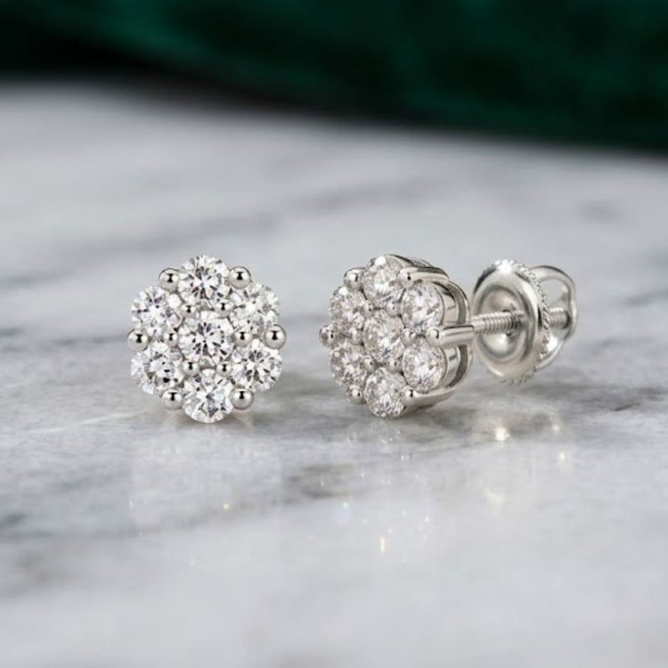A pair of 10k Gold Diamond Flower Cluster Earrings 1 Cttw rests on a gray marble surface, with one earring standing upright and the other lying on its side. Each stud features natural round diamonds in a dazzling floral cluster design.