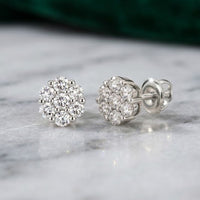 A pair of 10k Gold Diamond Flower Cluster Earrings 1 Cttw rests on a gray marble surface, with one earring standing upright and the other lying on its side. Each stud features natural round diamonds in a dazzling floral cluster design.