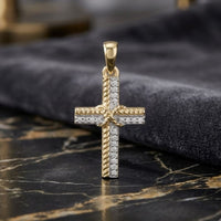 The 1/12CTW Natural Diamond Cross Pendant in 10K gold showcases a twisted rope edge and round center, accented with natural diamonds. The pendant is displayed on marble with a dark cloth backdrop.