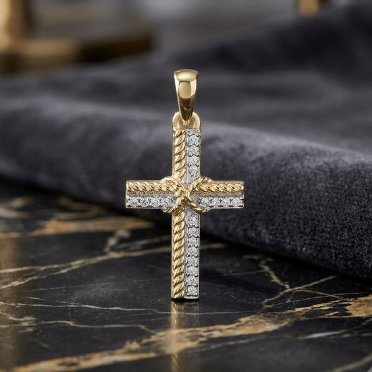 The 1/12CTW Natural Diamond Cross Pendant in 10K gold showcases a twisted rope edge and round center, accented with natural diamonds. The pendant is displayed on marble with a dark cloth backdrop.