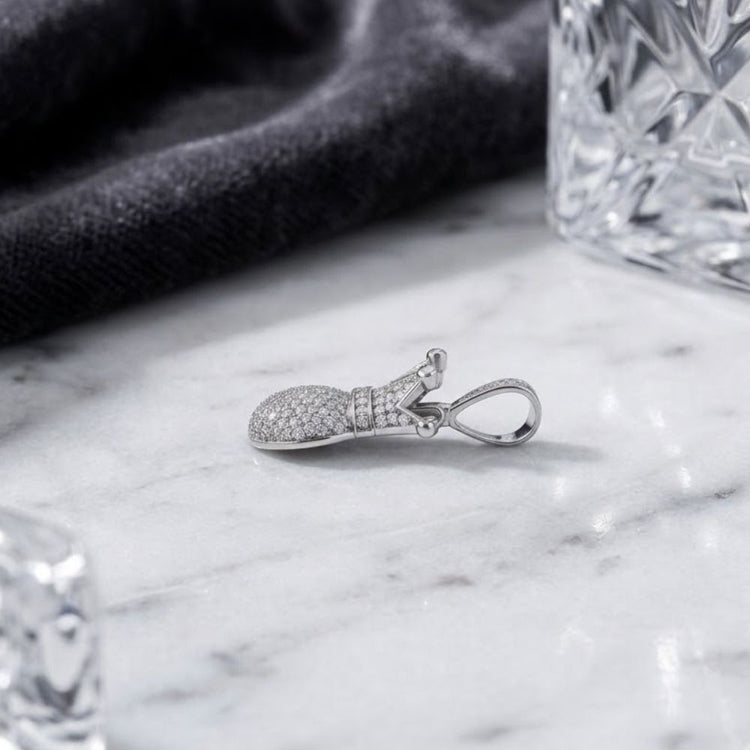 A gleaming 925 Sterling Silver Crown Heart Pendant, encrusted with petite diamonds, rests on a white marble surface beside a dark cloth and clear glass objects. This elegant pendant captures delicate detail and charm, making it an enchanting keepsake.