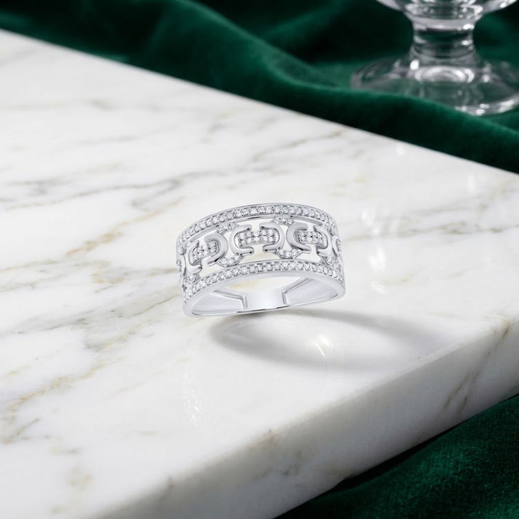 The 1/3 CT Natural Diamond Men’s Ring in Sterling Silver, featuring a geometric design and small embedded diamonds, is displayed on a white marble surface beside emerald green fabric and a glass stem.