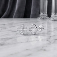 1/4 CTW Natural Diamond Clover Earrings in 10K Gold rest on a marble surface, with a blurred glass object in the background. These earrings feature a clover-inspired design adorned with round and baguette natural diamonds for an elegant touch.