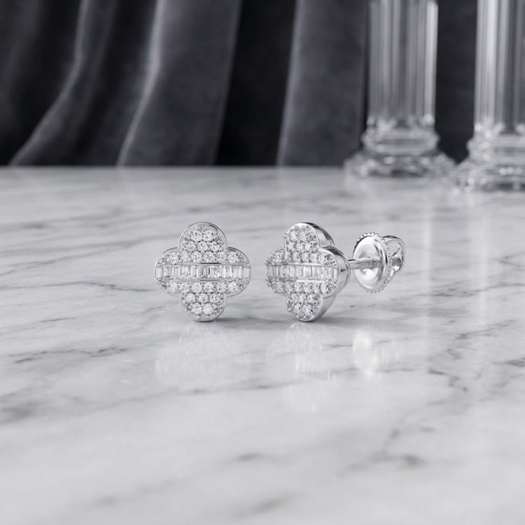 1/4 CTW Natural Diamond Clover Earrings in 10K Gold rest on a marble surface, with a blurred glass object in the background. These earrings feature a clover-inspired design adorned with round and baguette natural diamonds for an elegant touch.