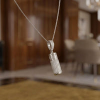 A close-up of the 10K Gold Diamond Urn Cylinder Pendant 1 CTTW hanging from a delicate chain, set against a softly blurred modern room in brown and beige tones.