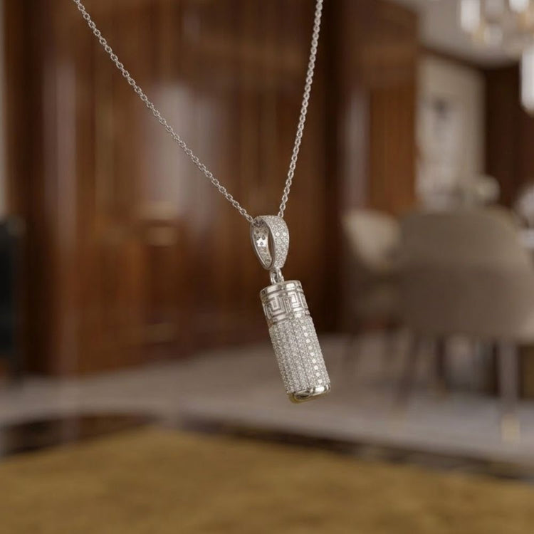 A close-up of the 10K Gold Diamond Urn Cylinder Pendant 1 CTTW hanging from a delicate chain, set against a softly blurred modern room in brown and beige tones.