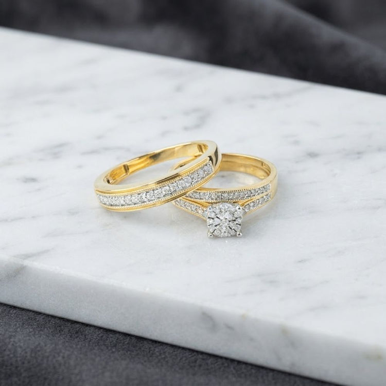 Two gold rings from the 3/8 CTW 10k Yellow Gold Wedding Trio Set rest on a white marble surface; one showcases a row of sparkling diamonds, while the other features a central round diamond accented by smaller stones along the band.