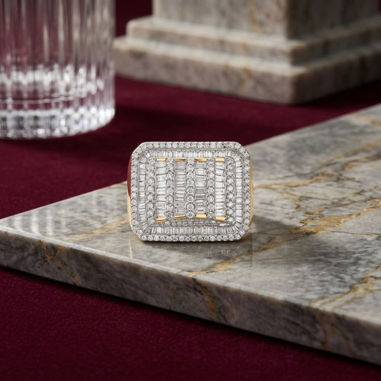 The 10k Yellow Gold Baguette Diamond Rectangle Ring, featuring multiple rows of baguette and round diamonds in a striking rectangle design, is beautifully displayed on a polished marble surface with a burgundy background and glassware in the distance.