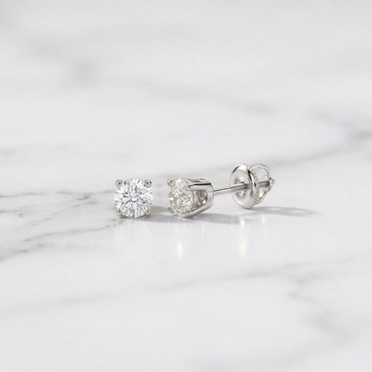A pair of 14k Gold Round Diamond Solitaire Stud Earrings 1/5 CTTW with sparkling diamonds in gold prong settings resting on a white marble surface.