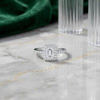 A stunning 14k Gold Emerald Diamond Halo Bridal Wedding Ring Set, featuring a square halo and pavé band, rests elegantly on a marble surface, accented by hints of green velvet fabric and glass decor in the background.