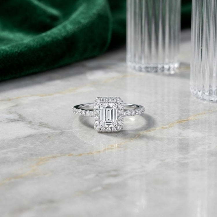 A stunning 14k Gold Emerald Diamond Halo Bridal Wedding Ring Set, featuring a square halo and pavé band, rests elegantly on a marble surface, accented by hints of green velvet fabric and glass decor in the background.