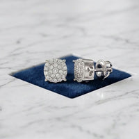 A pair of 10k Gold Diamond Cluster Earrings – 1/8 Cttw are displayed on a blue velvet square set into a white marble surface, showcasing their elegant cushion-shaped halos.