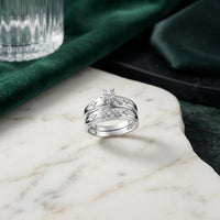 The 10k Gold Diamond Matching Wedding Ring Set, featuring a central diamond and intricately crisscrossed bands, is elegantly displayed on a white marble surface alongside complementary round diamond wedding bands. In the background, a softly draped green cloth and a sleek glass lend an air of subtle sophistication to the display.