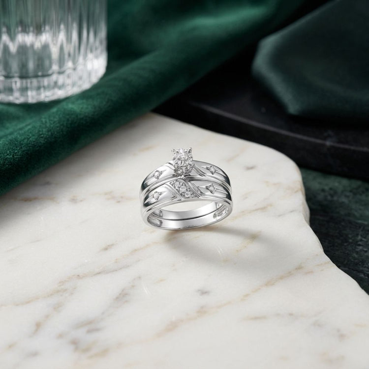 The 10k Gold Diamond Matching Wedding Ring Set, featuring a central diamond and intricately crisscrossed bands, is elegantly displayed on a white marble surface alongside complementary round diamond wedding bands. In the background, a softly draped green cloth and a sleek glass lend an air of subtle sophistication to the display.