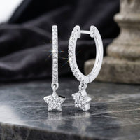 A pair of 10K Gold Diamond Star Hoop Earrings (1/5 CTTW) with delicate, sparkling star-shaped pendants, encrusted with clear diamonds, displayed on a marble surface with a stone column and black fabric in the background.
