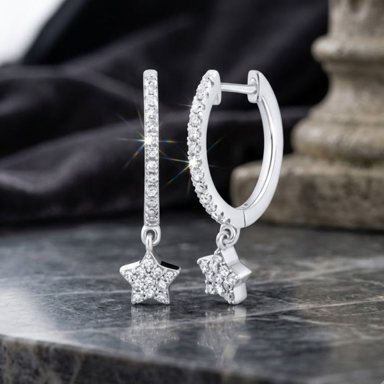 A pair of 10K Gold Diamond Star Hoop Earrings (1/5 CTTW) with delicate, sparkling star-shaped pendants, encrusted with clear diamonds, displayed on a marble surface with a stone column and black fabric in the background.
