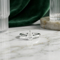 The 1/2CT Pear Diamond Solitaire Ring in 14k Gold is showcased on a white marble surface, complemented by glassware and green velvet fabric in the background.