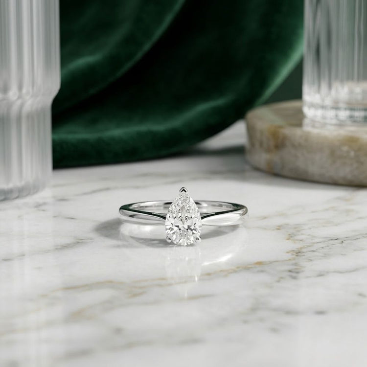 The 1/2CT Pear Diamond Solitaire Ring in 14k Gold is showcased on a white marble surface, complemented by glassware and green velvet fabric in the background.