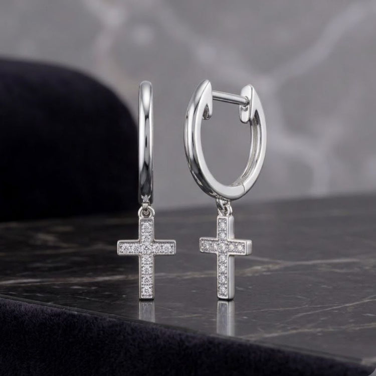 Two 10K yellow gold hoop dangle earrings, each adorned with a small, sparkling round diamond cross pendant (1/20CTW), rest on a black marble surface against a blurred gray background. Part of Nicole's Dream Collection, these elegant earrings exude timeless style and modern sophistication.