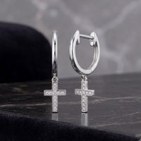 Two 10K yellow gold hoop dangle earrings, each adorned with a small, sparkling round diamond cross pendant (1/20CTW), rest on a black marble surface against a blurred gray background. Part of Nicole's Dream Collection, these elegant earrings exude timeless style and modern sophistication.