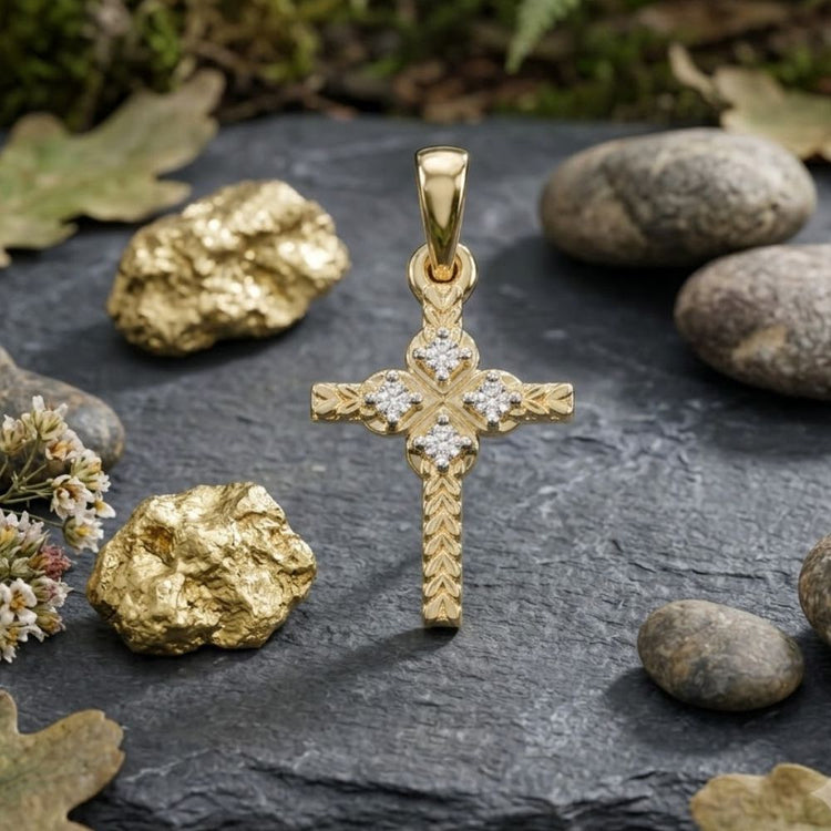 The 1/20CTW Natural Diamond Cross Pendant in 10K gold is shown on a dark stone slab, accented by gold nuggets, smooth pebbles, oak leaves, and small white flowers.