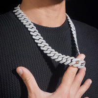 A person in a ribbed black sweater holds the Moissanite Cuban Links Chain 20mm - 925 Sterling Silver, a thick, shiny necklace adorned with sparkling stones, against their chest. The background is black.